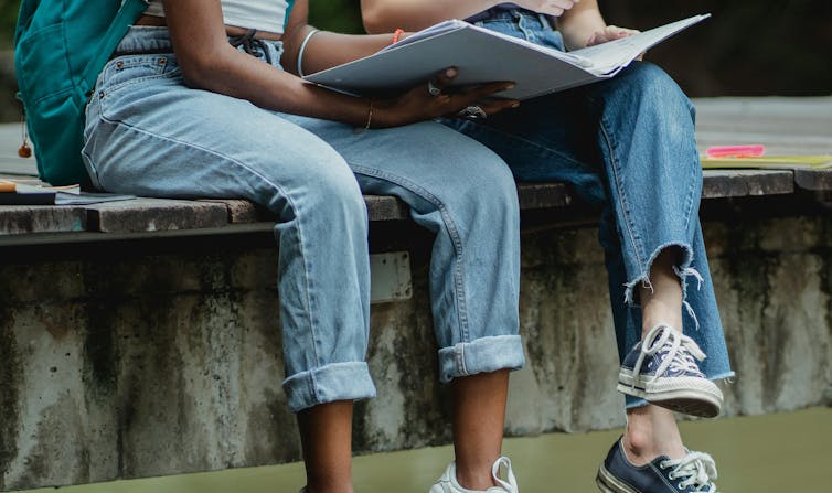 Two young people in jeans and sneakers sit on a low wall, with a folder.